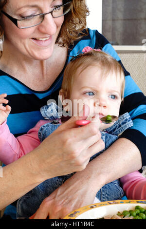 A Caucasian woman grandmother feeds her baby one year old girl grandaughter food from a plate. Kansas, USA. Stock Photo