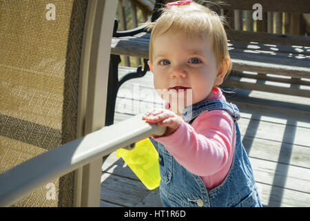 One year old smiling Caucasian baby girl in overalls with a drink cup, standing on an outdoor deck with sunshine. Kansas, USA. Stock Photo