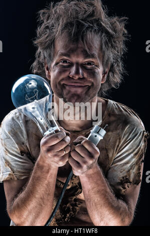 Portrait of burnt man with light bulb and electric plug over black background. Young man with dirty burnt face in funny sad expression in electricity  Stock Photo