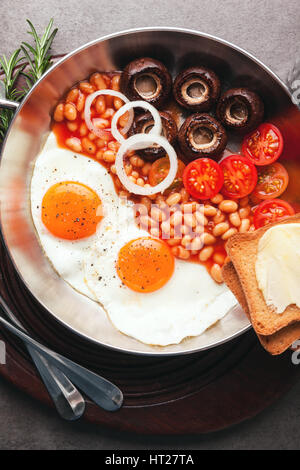 Breakfast with fried eggs in cast-iron pan, broken egg, tomatoes, on cutting board over grey stone background. Top view with copy space Stock Photo