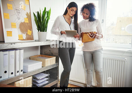 Two young entrepreneurs discussing packaging together as they stand working on a design or branding on a tablet for their small business, shelves of b Stock Photo