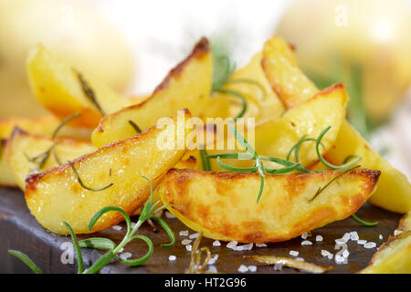 Baked potato wedges with rosemary served on a shabby cutting board with potatoes in the background Stock Photo