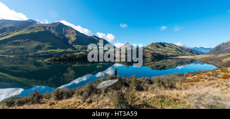 Hiker standing on rocks stretching out arms, mountains reflecting in lake, Moke Lake near Queenstown, Otago Region, Southland Stock Photo
