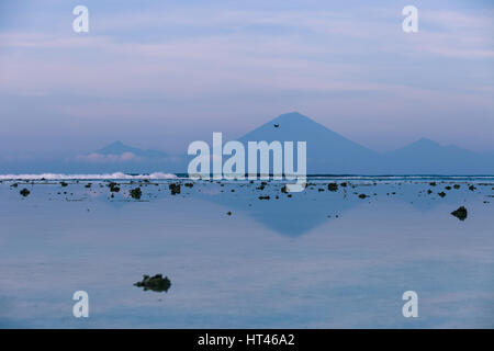 Views of the Agung volcano with reflection in the water off the island of Gili Trawangan in the early morning at low tide with exposed rocks, Indonesi Stock Photo