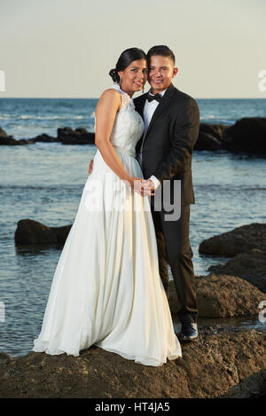 Wedding on ocean coast of hispanic couple. Young bride and groom stand on stone rock Stock Photo