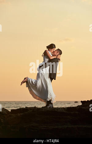 Groom hold and kiss bride on ocean sunset beach background. Married couple kissing on orange sunset sky Stock Photo
