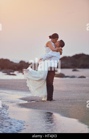 Happy kissing groom and bride stand on ocean coastline in sunset light. Exotic wedding on beach island with pink sky background Stock Photo