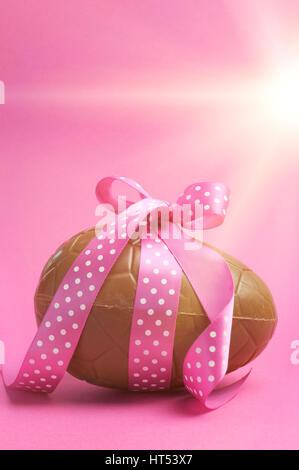 Large Happy Easter chocolate Easter egg with pink polka dot ribbon tied in a bow against a pretty feminine pink background, with faded lens flare filt Stock Photo