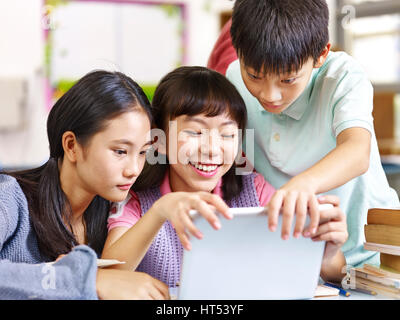 three happy asian elementary school students looking at tablet computer smiling in classroom. Stock Photo