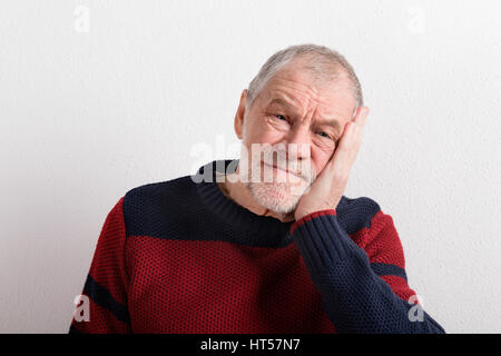 Upset senior man in red and blue sweater holding head. Studio shot against white wall. Stock Photo