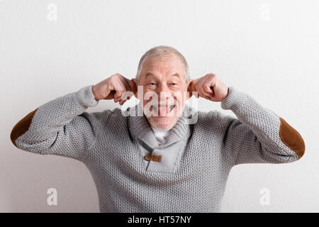 Handsome senior man in gray sweater smiling, sticking out tongue and pulling his ears. Studio shot against white wall. Stock Photo