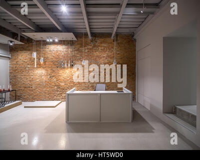 Luminous office in a loft style with brick and gray walls. There are many different hanging lamps. Also there is a gray reception desk with a laptop a Stock Photo