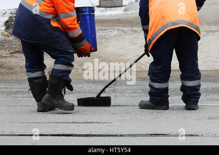 Two road workers in overalls are poured with hot tar on the edge of the road strip when repairing the road. Stock Photo