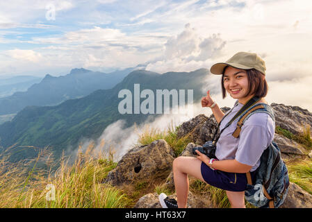 Happy hiker asian cute teens girl with dslr camera caps and backpack stand smiling poses thumb up on top of mountain at viewpoint Phu Chi Fa Stock Photo