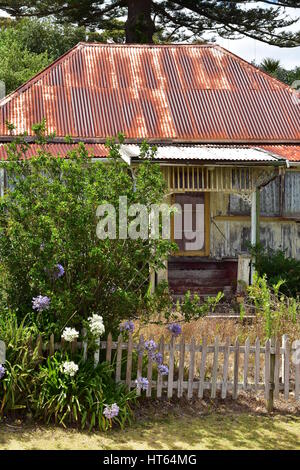 Weathered wooden house in very sand shape with roof of rusty corrugated sheet metal among greenery behind small wooden fence. Stock Photo