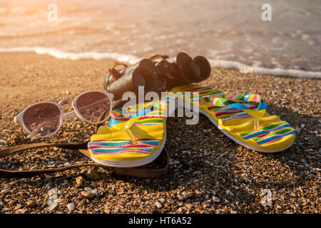 Flip flops and binoculars. Sunglasses are lying on sand. Explore new horizons. Be ready for summer adventures. Stock Photo
