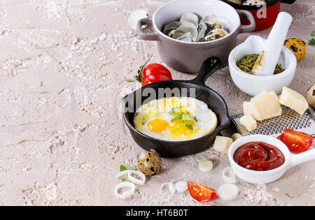 Breakfast with fried quail eggs in iron cast pan, cherry tomatoes, onion, ketchup sauce, seasonings in mortar, cheese grater, egg shell in pot. Beige  Stock Photo