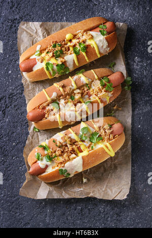 Hot dogs with sausage, fried onion, coriander leaves, cheese sauce and mustard, served on baking paper over black concrete texture background. Fast fo Stock Photo