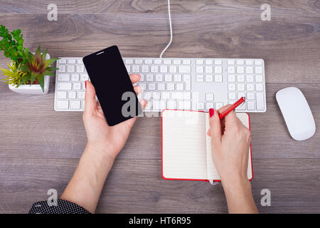 Closeup of female hand writing in a notebook and use a mobile phone on the desk with a keyboard Stock Photo