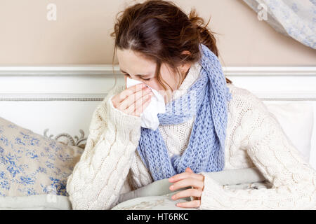 Caught a cold. Closeup image of young sick woman blowing her nose while sitting on bed. Stock Photo