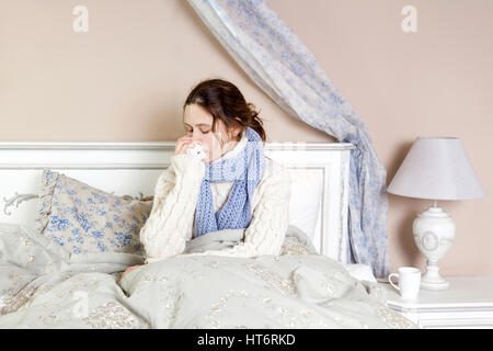 Flu. Closeup image of frustrated sick woman lying in bed in thick blue scarf holding tissue by her nose. Stock Photo