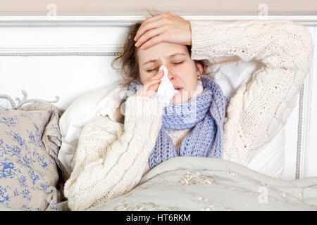 Flu. Closeup image of frustrated sick woman lying in bed in thick blue scarf holding tissue by her nose and touching her head with closed eyes. Stock Photo