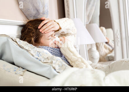 Flu. Closeup image of frustrated sick woman lying in bed in thick blue scarf holding tissue by her nose and touching her head with closed eyes. Stock Photo