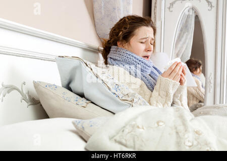 Sneezing beauty. Young sick woman with blue scarf on her neck sitting in bed and sneezing on her bed. Stock Photo