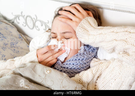 Flu. Closeup image of frustrated sick woman lying in bed in thick blue scarf holding tissue by her nose and touching her head and blowing her nose wit Stock Photo