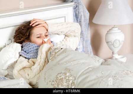 Flu. frustrated sick woman lying in bed in thick blue scarf holding tissue by her nose and touching her head. Stock Photo