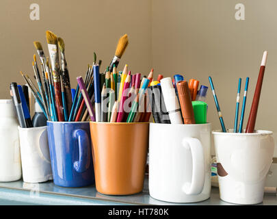 Close up of different used paint brushes, sharpened colored pencils, pens, and markers on colored mugs over blue table and beige background Stock Photo