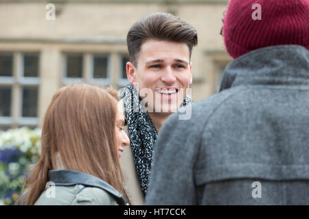A happy male student smiles as he stands amongst his friends outdoors. They are taking a break from university in the city centre. Stock Photo