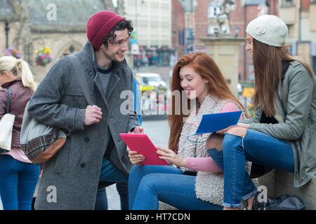 Three friends smile together as they take a break from university to study in the city centre. They are looking at their digital tablet and notebooks. Stock Photo