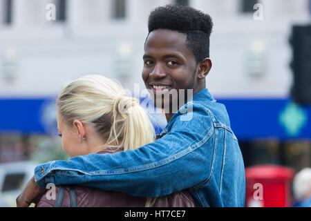 A happy young man smiles at the camera as he wraps his arm around his girlfriend's shoulder. She has her back to the camera. Stock Photo