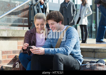 A happy young couple smile as they sit on some steps in the city looking at a mobile phone. There is a group of other students out of focus in the bac Stock Photo