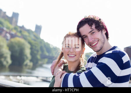 A happy young couple smile as they relax outdoors by the river. They are in an embrace. Stock Photo