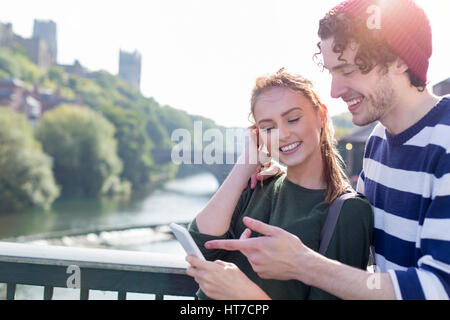 A happy young couple smile as they relax outdoors by the river. They are using a mobile phone laughing together. Stock Photo