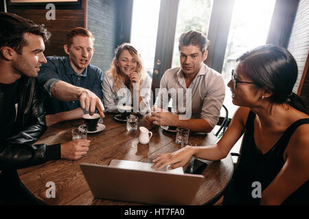 Group of young friends sitting at cafe with laptop and discussing. Young men and women looking at laptop at coffee shop. Stock Photo