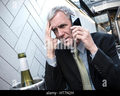 Businessman bad news stressed and worried on his iPhone 7 smartphone at alfresco café restaurant table with wine bottle in foreground Stock Photo