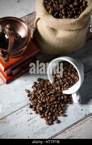 Coffee grain spill from a cup. Jute bag of roasted beans and mill in background Stock Photo