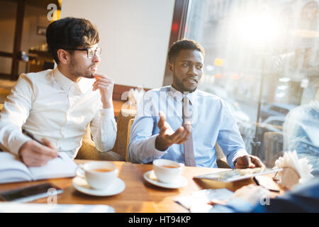 Confident African-American manager offering new company strategies to his intent bearded partner in glasses while having tea break in cafe. Stock Photo
