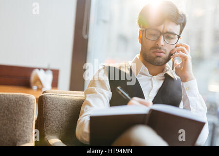 Portrait of busy dark-haired bearded executive in eyeglasses and waistcoat sitting in cafe, talking on phone and writing down necessary business data  Stock Photo