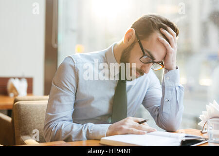 Bearded pensive businessman in shirt and tie sitting in cafe bent over notepad, holding head with his hand and thinking hard over solving company prob Stock Photo