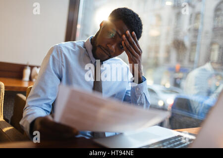 Focused African-American office manager sitting at cafe table with laptop, reading important documents with puzzled expression and holding head with h Stock Photo