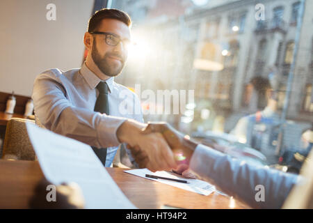 Confident bearded businessman in eyeglasses sitting at cafe table and shaking hand of his partner and smiling after making profitable agreement. Stock Photo