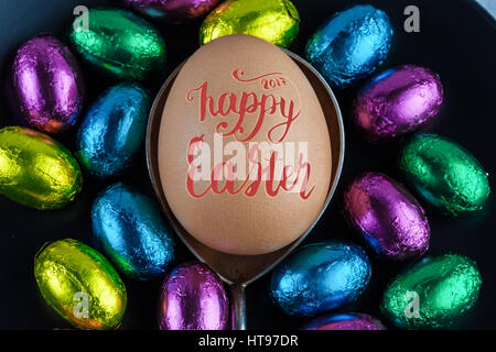 Happy Easter 2017 lettering written on Easter Egg placed on silver spoon with small chocolate eggs in colorful foil for decoration Stock Photo