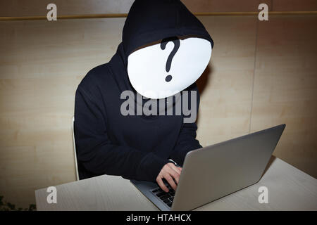 Anonymous hacker in black hoodie hiding his face under white mask with question mark and sitting at desk with laptop Stock Photo