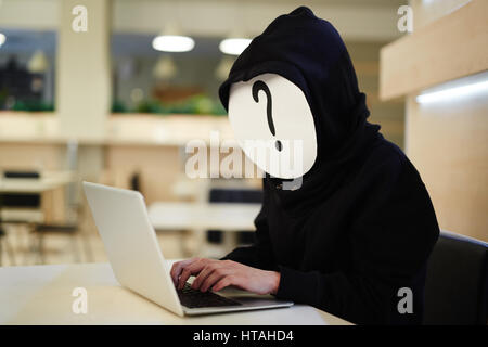 Internet security concept: anonymous hacker in question mark mask and black hoodie using laptop while sitting in cafe Stock Photo