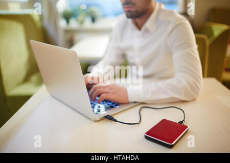 Serious bearded journalist sitting in PC cafe and writing article for magazine on laptop connected to external hard drive Stock Photo