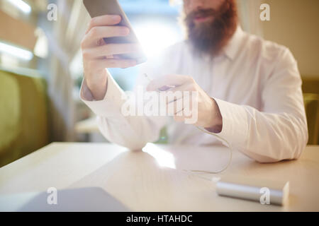 Process of charging smartphone: bearded businessman in buttoned-up white shirt holding device in one hand and cable of power bank in another while sit Stock Photo